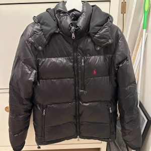 Svart pufferjacka från Polo Ralph Lauren - Svart pufferjacka från Polo Ralph Lauren med röd logga på bröstet. Minimal defekt (SE PÅ SIDA 4) Jackan har avtagbar huva, flera dragkedjefickor och quiltad design. Materialet är glansig polyester och jackan är långärmad med normal passform. Perfekt för kalla dagar.