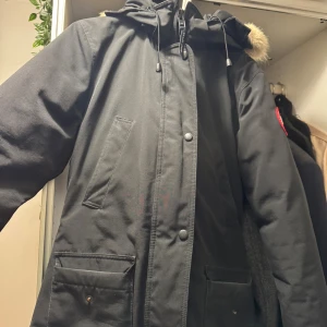 Svart parkas från Canada Goose modell trillium - Säljer en svart parkas från Canada Goose med huva och fluffig päls. Jackan har dragkedja, knappar och stora fickor framtill. Storleken är S, passar också M då man kan justera midjan. Jackan är i bra skick och inget slitage eller liknande finns.