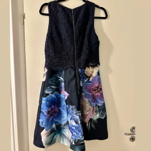 Mörkblå spetsklänning med blommigt mönster  - Supersnygg mörkblå klänning med överdel i spets och kjol med stora blommor i blått, lila och vitt. Klänningen är ärmlös och har en dragkedja bak. Kjolen är utsvängd och ger en riktigt fin siluett. Perfekt för dig som vill sticka ut med ett färgstarkt blommönster.