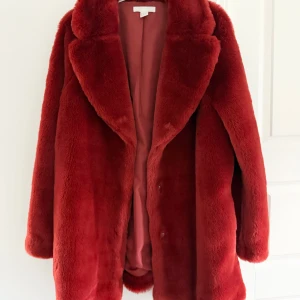 RostRöd pälsjacka - Säljer en fluffig roströd pälsjacka med bred krage och helröd insida. Den är tillverkad i mjukt fuskpälsmaterial och har knäppning framtill. Aldrig använd.