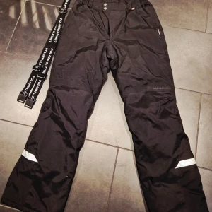 Svarta täckbyxor Polarn O. Pyret - Nya använda 1 gång .Svarta täckbyxor från Polarn O. Pyret med avtagbara hängslen, reflexdetaljer och knäppning i midjan. Vattentäta, vindtäta och andasmaterial med återvunnen vaddering. Perfekta för kalla dagar och vinteraktiviteter.