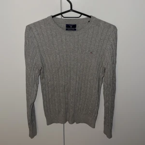 Grå kabelstickad tröja från Gant - Kabelstickad grå tröja från Gant i stretchig bomull. Klassisk rund halsringning och diskret broderad logga på bröstet. Långärmad modell med ribbade muddar och en clean, tidlös look som funkar till allt.