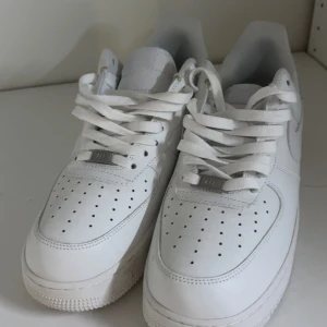 Nike Air Force 1 helvita sneakers - Nike Air Force 1 sneakers i helvitt med klassisk design och perforerad tå. Skorna har snörning, platt sula och är tillverkade i skinn med en rund tå. Swoosh-loggan syns på sidan och sulan har det ikoniska cirkelmönstret. Perfekta för dig som gillar cleana och tidlösa sneakers.