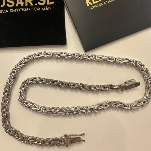 Silverfärgat halsband från Kejsar.se - Kejsarlänk i silver från Kejsar.se med ett flätat och detaljerat kedjemönster. Perfekt för dig som gillar exklusiva och stilrena smycken med lite tyngd. Låset är diskret och smidigt, och halsbandet kommer med originalpåse. (55cm) lång