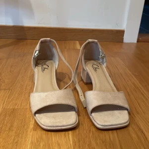 Beige sandaler med blockklack - Snygga sandaler från Glam i beige mocka med bred rem över tårna och justerbart spänne vid ankeln. Skorna har en fyrkantig tå och stabil blockklack som ger extra komfort. Perfekt för dig som gillar stilrena och enkla detaljer.