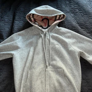 Grå hoodie från Burberry med dragkedja - Säljer en grå hoodie med klassiskt rutigt foder i huvan. Tröjan har dragkedja framtill, snörning vid huvan och två fickor. Perfekt för dig som gillar stilrena och exklusiva plagg med en twist.