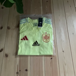 Spaniens gulblå fotbollströja Adidas S - Grym fotbollströja från Adidas i slim fit, officiell Spanien-variant. Tröjan är ljusgul med blå detaljer och röda inslag, har korta ärmar och V-ringning. Materialet är lätt polyester med ventilerande paneler. Adidas-logga och Spaniens emblem på bröstet.