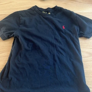 Svart t-shirt från Polo Ralph Lauren - Klassisk svart t-shirt från Polo Ralph Lauren med röd broderad logga på bröstet. T-shirten har rund halsringning och korta ärmar. Tillverkad i mjuk bomull för en skön känsla. Perfekt för en stilren och avslappnad look.