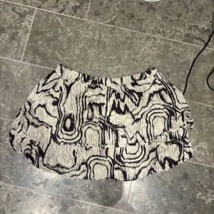 Svartvit kortkjol med abstrakt mönster - Kort kjol i svart och vitt med ett unikt abstrakt mönster som påminner om träådring. Kjolen har en lätt volang och resår i midjan för skön passform. Perfekt för dig som vill sticka ut med en cool och annorlunda stil.