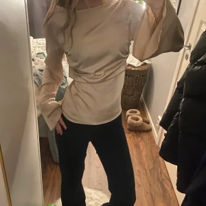 Champagnefärgad satintopp med vida ärmar - Säljer en champagnefärgad satintopp med långa, vida trumpetärmar. Toppen har en slät, glansig yta och en figurnära passform som ger en elegant look. Perfekt för dig som vill sticka ut med en unik och trendig topp.