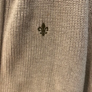 Beige kofta med dragkedja från Morris - Ribbstickad beige kofta från Morris i storlek XS. Koftan har hel dragkedja framtill, hög krage och diskret broderad logga på bröstet. Perfekt för lager-på-lager och snygg till jeans eller kjol.