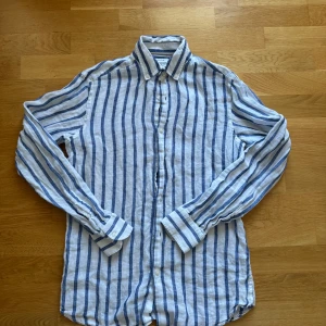 Randig linneskjorta från Massimo Dutti - Snygg randig skjorta från Massimo Dutti i 100% linne. Skjortan är vit med blå vertikala ränder, har klassisk krage och knäppning framtill. Perfekt för en fräsch och avslappnad stil under varmare dagar.