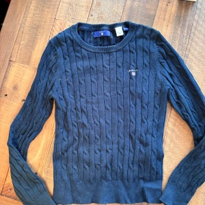 Mörkblå stickad tröja från GANT - Snygg mörkblå stickad tröja från GANT med klassiskt kabelstickat mönster och rund hals. Tröjan har långärmade ärmar och en liten broderad logga på bröstet. Perfekt för dig som gillar stilrena och tidlösa plagg. 134/140 