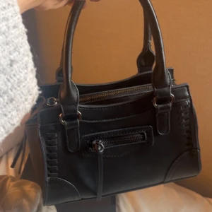 Gina väska  - Snygg svart handväska i skinnimitation med två stadiga handtag och dekorativa sömmar. Väskan har en dragkedja upptill och en mindre ficka med dragkedja framtill, perfekt för småprylar. Stilren och enkel design som passar till många outfits.