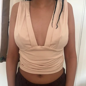 Beige croppad topp med djup v-ringning - Säljer en beige croppad topp med breda axelband och djup v-ringning både fram och bak. Toppen har en figurnära passform och är tillverkad i ett mjukt, stretchigt material som sitter snyggt på kroppen. Passar bra till någon middag eller fest💕har använt den typ en gång
