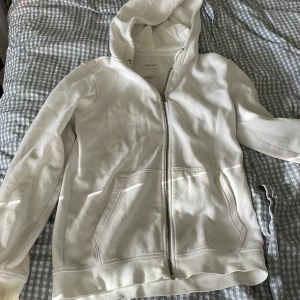 Vit hoodie med dragkedja från Pier One - Vit hoodie från Pier One med dragkedja framtill och två fickor. Klassisk modell med huva och långa ärmar. Tillverkad i mjuk bomull, perfekt för chill dagar eller när du vill ha en enkel och clean look. Lite defekter som man kan se på bilderna