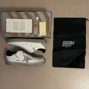 GGDB Superstar vita sneakers med grå stjärna - Säljer ett par helt oanvända GGDB Superstar sneakers i vitt skinn med grå mockadetaljer och ikonisk stjärna på sidan. Skorna har grå snörning, svart häl och slitna detaljer för en cool vintage-look. Kommer med originalkartong och dustbag.