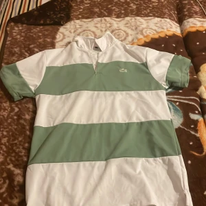 Randig Lacoste t-shirt i grönt och vitt - Snygg t-shirt från Lacoste med breda gröna och vita ränder. Klassisk krage med liten öppning framtill och det ikoniska krokodilmärket på bröstet. Perfekt för dig som gillar sportig och stilren look.