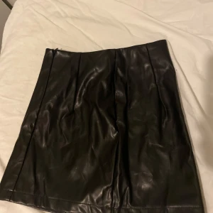 Svart skinnkjol med slits - Snygg svart kort skinnkjol i syntetmaterial med stilren design och diskret slits framtill. Kjolen har en rak passform och är perfekt för dig som gillar en edgy look. Passar till många olika stilar och är lätt att matcha med både boots och sneakers.