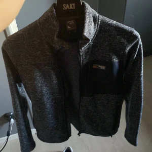 Mörkgrå fleecekofta från Sprayway - Mysig mörkgrå fleecekofta från Sprayway med hel dragkedja och hög krage. Tröjan har en bröstficka med dragkedja och diskret logga på bröstet. Perfekt för kyliga dagar och enkel att matcha med andra plagg.