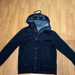 Mörkblå hoodiejacka från C.P. Company - Säljer en mörkblå hoodiejacka från C.P. Company med ikoniska glasögon i huvan. Jackan har knappar framtill, två stora fickor och är tillverkad i bomull och polyester. L men passar mig i M