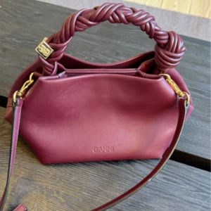 Vinröd handväska bou Ganni - Säljer en vinröd handväska från Ganni med flätat handtag och justerbar axelrem. Väskan är tillverkad i slätt skinn och har guldfärgade metalldetaljer. I modellen Bou