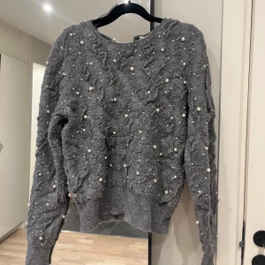 Grå stickad tröja med pärlor från Zara - Mysig grå stickad tröja från Zara med cool struktur och pärldetaljer över hela tröjan. Långärmad modell med ribbade muddar och avslappnad passform. Perfekt för dig som vill ha något extra till din outfit.