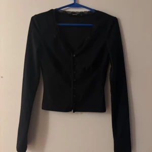 Svart långärmad blus från BikBok - Snygg svart blus från BikBok i storlek XS. Modellen är figurnära med långa ärmar, knappar framtill och v-ringning med spetsdetalj vid halsen. Perfekt att styla med jeans eller kjol för en trendig look.