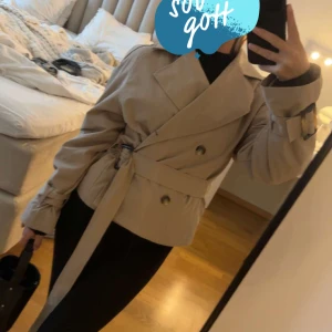 Kort beige trenchcoat med bälte - Snygg kort trenchcoat i beige med dubbelknäppning, bälte i midjan och spänndetaljer vid ärmsluten. Jackan har en klassisk krage och en loose passform som ger en cool och avslappnad look. storlek xxs/xs