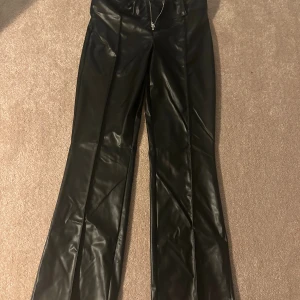 Svarta bootcut byxor i skinnimitation från Gina Tricot - Snygga svarta byxor från Gina Tricot i skinnimitation med bootcut passform. Byxorna har dragkedja framtill, bälteshällor och en stilren look som passar perfekt till en trendig outfit.