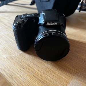 Nikon Bridgekamera med väska - Snygg Nikon bridgekamera i svart, säljs med praktisk axelväska med fack för minneskort. Kameran och väska ser ut att vara i gott skick utan synligt slitage. Perfekt för dig som vill ta bättre bilder och ha allt samlat och skyddat.