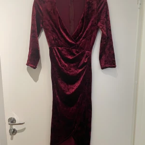 Vinröd sammetsklänning Petite - Vinröd långklänning i mjuk sammet med omlott framtill och djup v-ringning. Klänningen har trekvartsärmar och en dold dragkedja i ryggen. Snyggt figurnära passform och elegant fall. Perfekt för dig som vill sticka ut med en lyxig känsla. Endast använd en gång. 