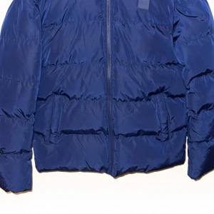 Blå pufferjacka från Urban Classics - Blå pufferjacka från Urban Classics med huva och dragkedja framtill. Jackan har en klassisk quiltad look, två stora fickor och en stilren logotyp på bröstet. Perfekt för kalla dagar när du vill hålla stilen och värmen.