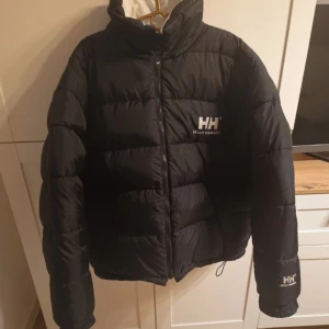 Svart pufferjacka från Helly Hansen - PASSA PÅ ! Svart pufferjacka från Helly Hansen med broderad logga på bröstet, ärmen och kragen. Jackan har hög krage, dragkedja framtill och elastiska muddar. Perfekt för kalla dagar och riktigt snygg streetstil.