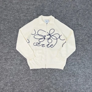 Arte Full Zip M - Säljer en krämvit kofta från Arte med hel dragkedja och snyggt blått grafiskt mönster över bröstet och ryggen. Tröjan är stickad i mjuk bomull, har ribbade muddar och två fickor framtill. Perfekt för dig som gillar stilrena men unika plagg.