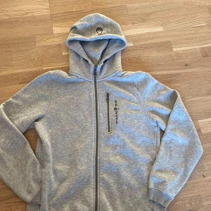Grå hoodie från Sail Racing - Snygg ljusgrå hoodie från Sail Racing med dragkedja framtill och bröstficka med zip. Tröjan har broderad logga på bröstet och huva med broderad detalj. Tillverkad i mjuk bomullsblandning och har långa ärmar med muddar.