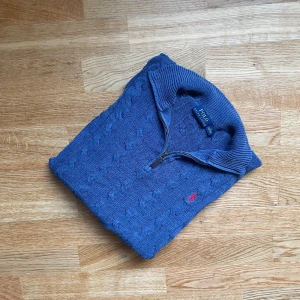 Blå kabelstickad half zip från Polo Ralph Lauren - Kabelstickad blå tröja från Polo Ralph Lauren med halv dragkedja och ribbade muddar. Klassisk röd broderad logga på bröstet. Tröjan har hög krage och är långärmad, perfekt för lager-på-lager under kyliga dagar.