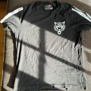Svart t-shirt från Plein Sport M - Svart t-shirt från Plein Sport med vit tigerlogga på bröstet och vita detaljer på axlarna. Klassisk rund hals och korta ärmar. Tillverkad i mjuk bomull med lite stretch för skön passform. Perfekt för dig som gillar sportig och stilren look.