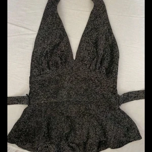 Glittrig svart omlott halterneck topp - Snygg svart glittrig halterneck topp med omlott och bred knytning i midjan. Toppen har djup v-ringning och volangliknande nederkant som ger en cool siluett. Perfekt för dig som vill sticka ut med lite extra glitter.