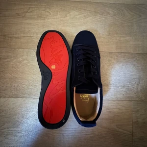 Christian Louboutin svarta sneakers - Snygga mörkblå sneakers från Christian Louboutin med klassisk röd sula och diskret broderad logga på hälen. Ovandel i mocka och insida i ljust skinn. Skorna har rund tå, platt sula och snörning framtill. Perfekta för dig som gillar exklusiva detaljer och stilren design.
