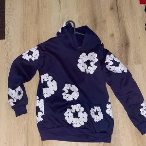 Blå hoodie från Denim Tears med blommor - Säljer en blå hoodie från Denim Tears med stora vita blommönster över hela tröjan. Tröjan har huva, långa ärmar och ribbade muddar. Perfekt för dig som vill sticka ut med en unik och trendig stil.