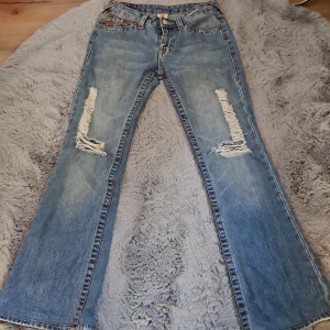 True Religion bootcut jeans - Snygga ljusblå bootcut jeans från True Religion med slitna detaljer på låren och hål i knäna med ikoniska broderade bakfickor. Klassisk femficksmodell med orange kontrastsömmar och låg midja. Mått i bild 3&4. Skriv om du har frågor