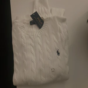 Vit kabelstickad tröja Polo Ralph Lauren - Vit kabelstickad tröja från Polo Ralph Lauren med klassisk rund halsringning och broderad marinblå logga på bröstet. Tröjan har långa ärmar och ribbade muddar för en snygg och tidlös look. Perfekt för dig som gillar stilrena plagg. Pris går att diskuteras 