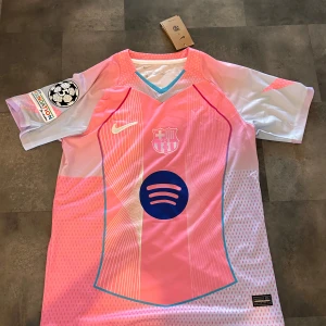 FC Barcelona Raphinha 11 rosa matchtröja - Snygg FC Barcelona fotbollströja från Nike i rosa och ljusblått med vita detaljer. Äkta. Tröjan har korta ärmar, V-ringning med blå kant, Champions League-märke på ärmen och tryck med Raphinha 11 på ryggen. Materialet är lätt och andas, perfekt för match eller träning. Pris kan diskuteras 
