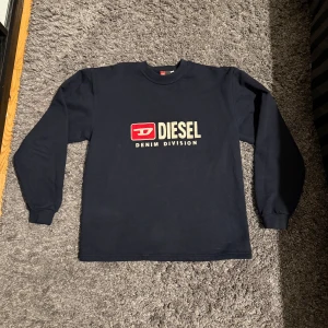 Svart långärmad tröja från Diesel - Svart långärmad tröja från Diesel med stort tryck på bröstet i vitt och rött där det står 'Diesel Denim Division'. Klassisk passform med rund hals och muddar vid ärmslut. Perfekt för dig som gillar streetwear och vill ha en enkel men snygg look.