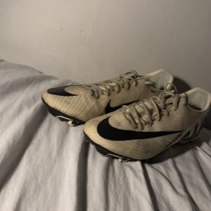 Nike Mercurial fotbollsskor beige/svart - Säljer ett par Nike Mercurial fotbollsskor i beige med svarta detaljer och stor Nike-logga på sidan. Skorna har snörning och är tillverkade i syntetmaterial, designade för att ge bra grepp på planen. Perfekta för dig som vill sticka ut med stil på matchen.