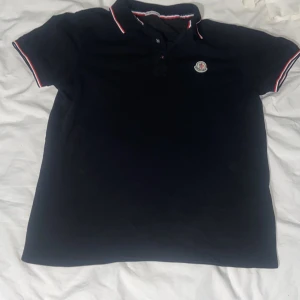 Svart Moncler pikétröja med röd/vit kant - Svart pikétröja från Moncler med klassisk krage och knappar framtill. Tröjan har Moncler-logga på bröstet och snygga rödvita detaljer på krage och ärmslut. Perfekt för en clean och sportig look.