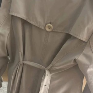 Beige trenchcoat med leopardfoder - Stilren beige trenchcoat från Tiger of Sweden med dubbelknäppning, stora knappar och bälte i midjan. Insidan har ett fett leopardmönstrat foder som ger jackan extra edge. Perfekt för dig som vill sticka ut med detaljer men ändå hålla det klassiskt.