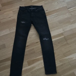 Svarta skinny jeans från Jack & Jones - Svarta skinny jeans från Jack & Jones med slitna detaljer vid knäna. Klassisk femficksmodell med knapp och dragkedja. Jeansen har en tight passform och är tillverkade i stretchigt denim för extra komfort.