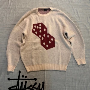 Beige Stüssy tröja med tärningsmotiv - Säljer en beige stickad tröja från Stüssy med stort rött tärningsmönster framtill. Tröjan har rund halsringning, ribbade muddar och är tillverkad i 100% bomull. Perfekt för dig som gillar streetwear och unika prints.
